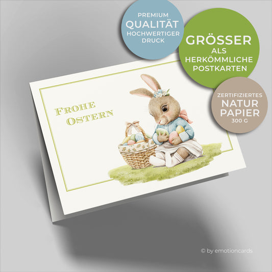 Osterkarte mit feinem grünen Doppelrahmen und kleinem Hasen im Gras sitzend mit Ostereiern und Osterkorb daneben stehend.