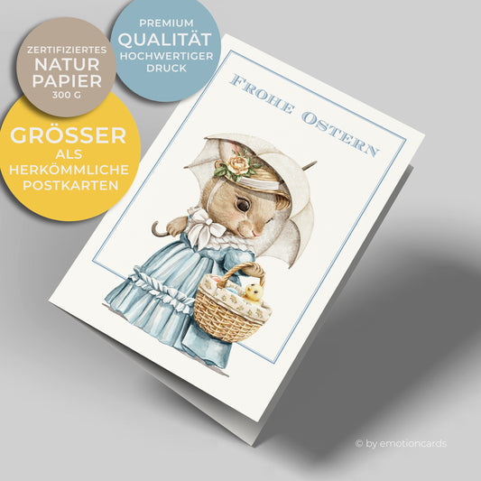 Osterkarte mit hellblauem feinen Doppelrahmen. Häsin mit Hut und Sonnenschirm in hellblauem Kleid trägt Osterkorb mit Eiern und kleinem gelben Küken darin.