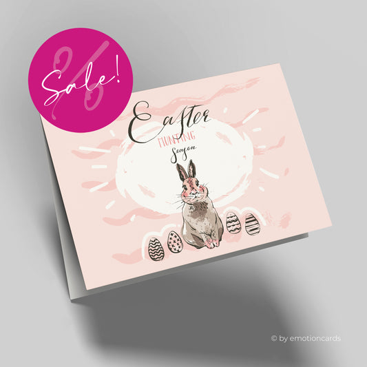 Moderne Osterkarte in Hellrosa mit braunem Osterhasen und 4 Ostereiern neben dem Hasen stehend. Englische Karte mit dem Spruch Easter Hunting Season