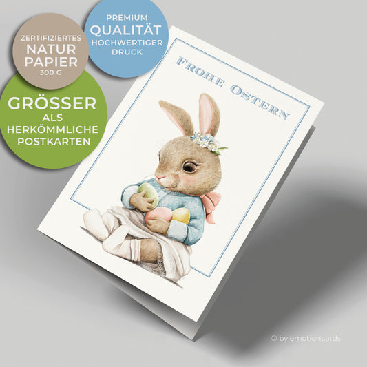 Süße Osterkarte im Hochformat mit feinem blauen Doppelrahmen. Kleine Häsin am Boden sitzend, die drei bunte Ostereier hält.